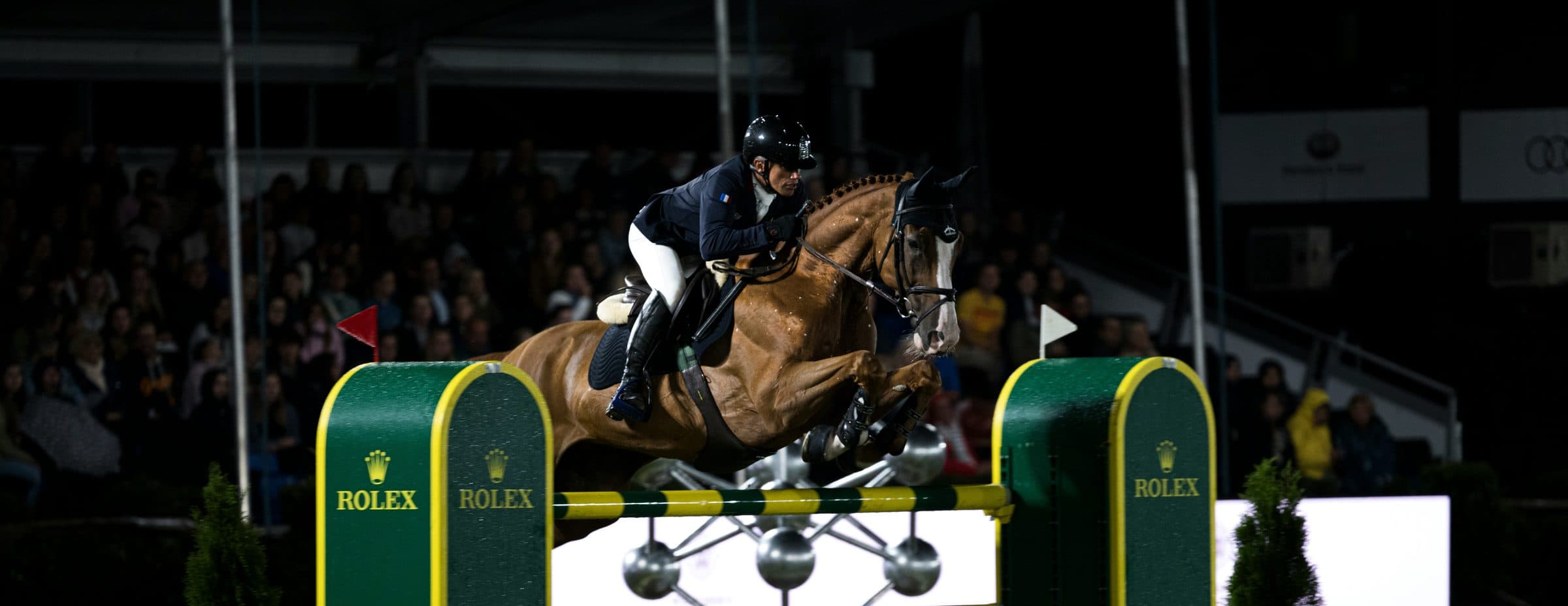 A horse and rider clearing a floodlit oxer in front of a packed grandstand